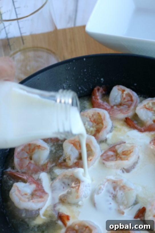 Rich heavy cream being poured into the skillet with cooking shrimp and garlic, beginning the transformation into a decadent creamy garlic butter sauce.