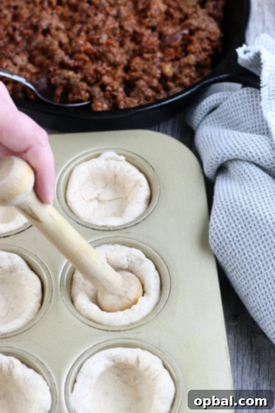 Cheeseburger Biscuit Poppers 8 A pair of hands gently pressing refrigerated biscuit dough into the cups of a greased muffin tin, forming the perfect base for the cheeseburger bites.