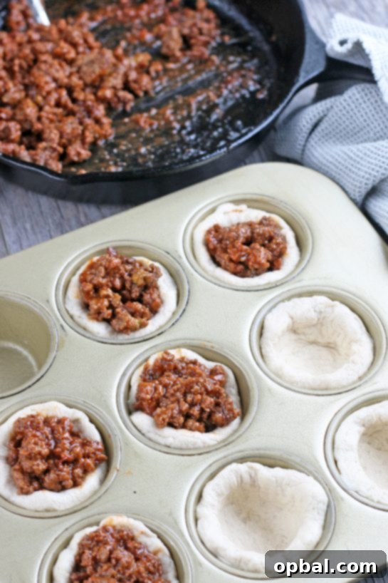 Cheeseburger Biscuit Poppers 9 The biscuit cups are now formed in the muffin tin, and a spoon is adding the flavorful ground beef mixture into each cup, filling them perfectly for baking.
