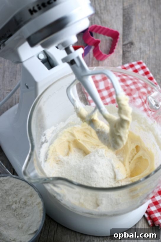 Crafting Edible Love Notes: Decorating Valentine's Conversation Heart Cookies 9 A stream of dry ingredients, specifically flour mixture, being gradually incorporated into the stand mixer bowl, ensuring a smooth and thoroughly combined batter for our Valentine's cookies.