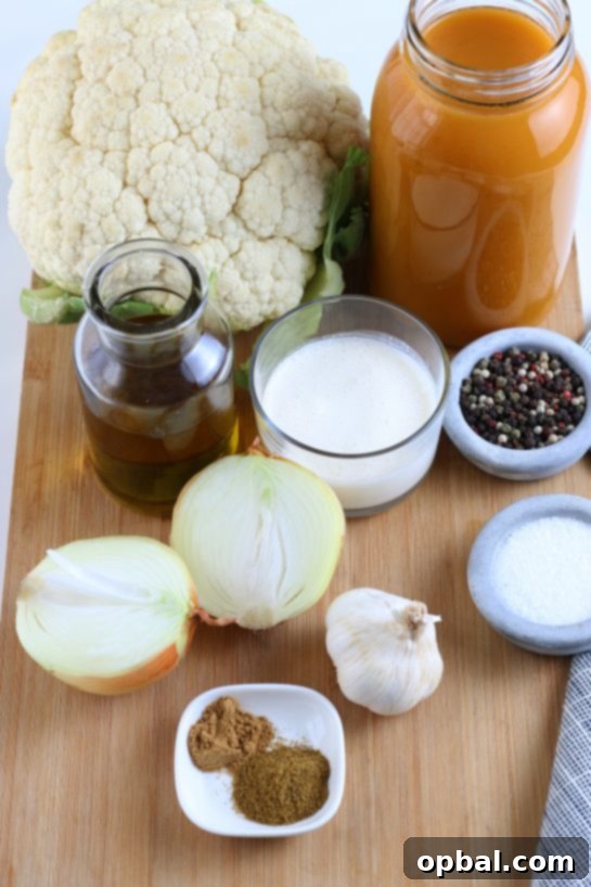All the fresh ingredients laid out on a kitchen counter, ready for cooking: a head of cauliflower, onion, garlic, olive oil, vegetable broth, heavy cream, cumin, coriander, salt, and pepper for the roasted cauliflower soup.