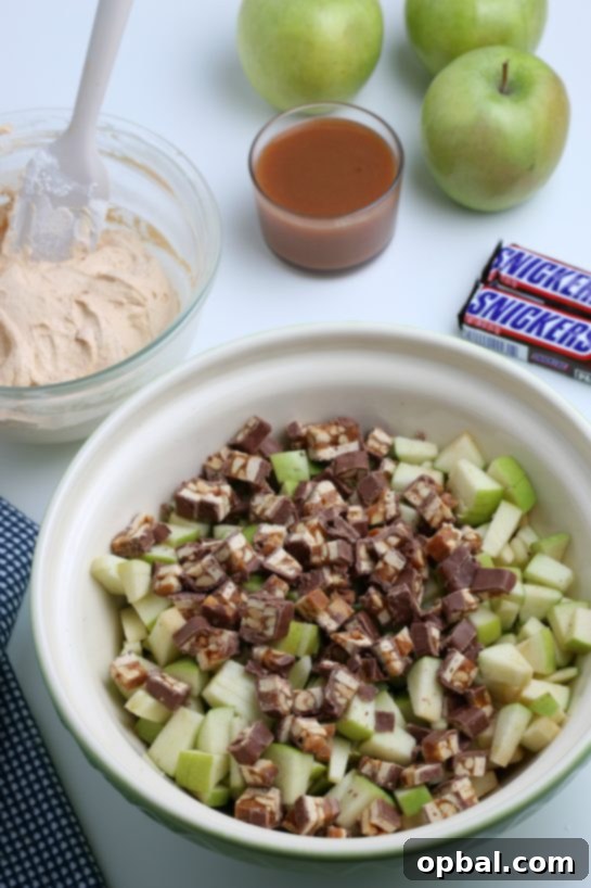 Caramel Apple Snickers Delight 5 Chopped Snickers candy bars are gently folded into a bowl with diced green apples, creating a delicious mix for the Caramel Apple Snickers Salad.