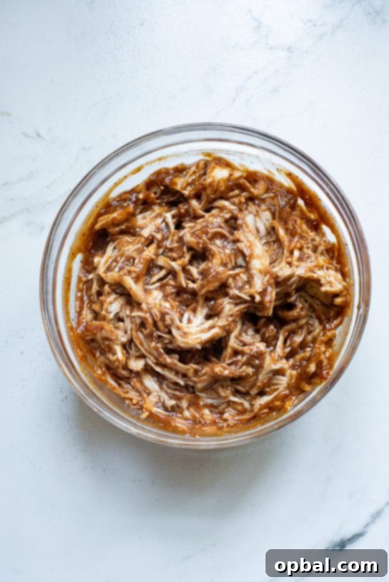 Shredded chicken being mixed with BBQ sauce and cayenne pepper in a bowl.